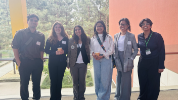 Six students dressed in semi-formal attire smile together during a conference, standing on a balcony with trees and a red-orange building wall behind them.